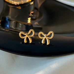 Minimal gold ribbon bow stud earrings on dark surface